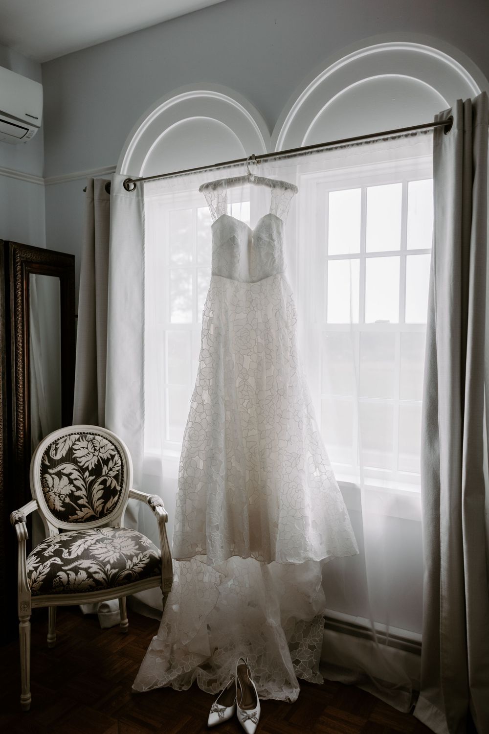 Strapless lace A-line wedding gown hanging in arched window with bridal heels below on hardwood floor.