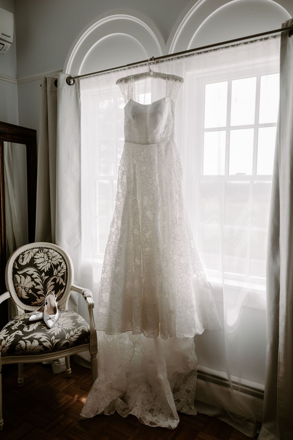 Elegant strapless lace wedding gown hanging in a sunlit arched window with bridal shoes on chair.