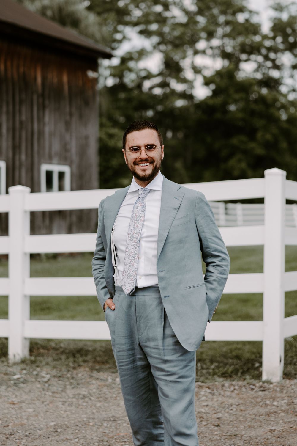 Smiling groom in a light blue suit with floral tie standing near a white fence and barn.