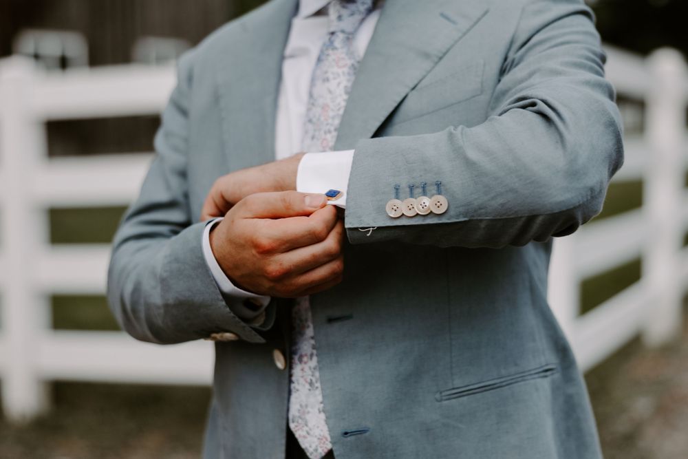Groom in light blue suit adjusting silver cufflinks on his sleeve near a white fence.
