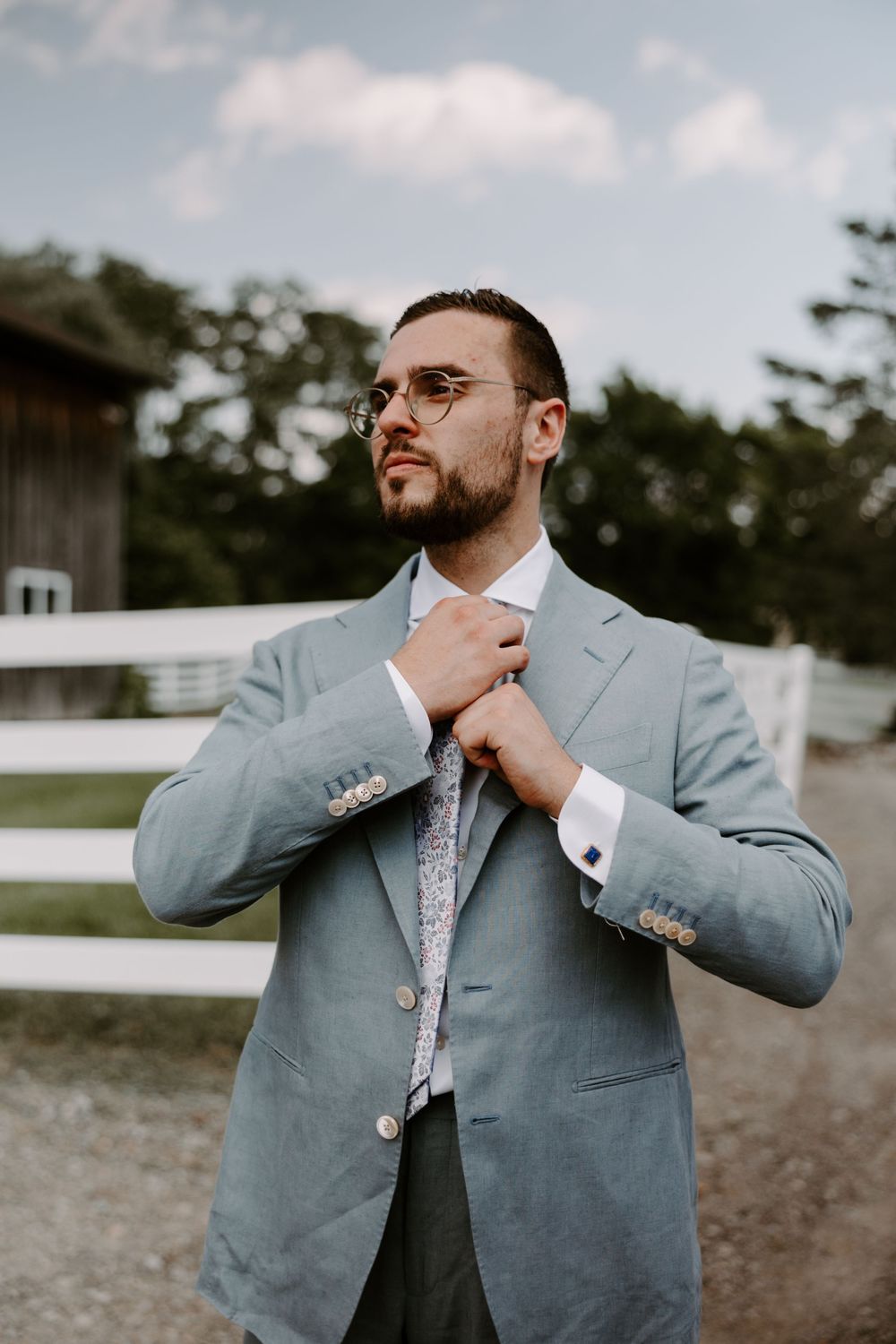 Groom in light blue suit adjusting floral tie outdoors near white fence.