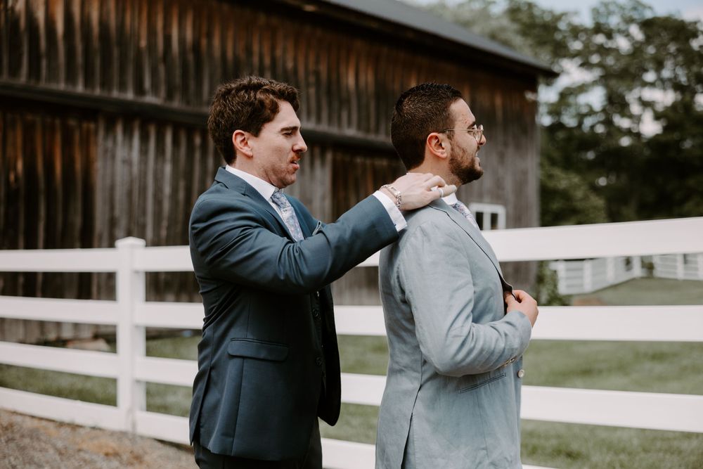 Groomsman helping groom adjust suit jacket near rustic barn and white fence.