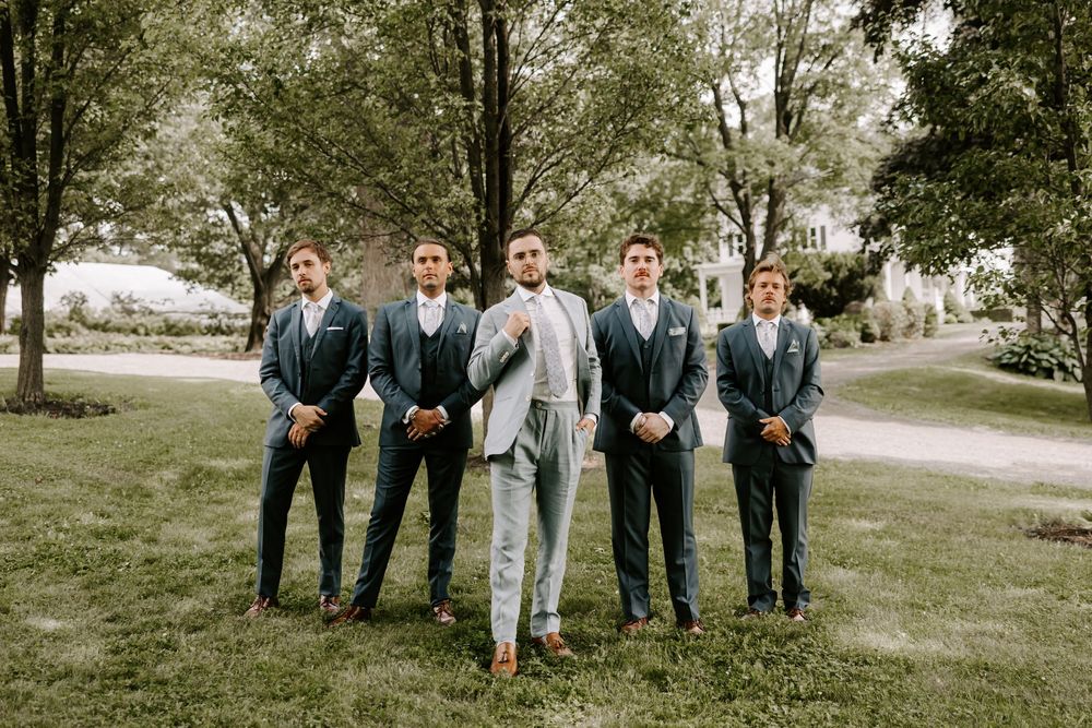 Groom in light gray suit standing with four groomsmen in navy suits on a green lawn.