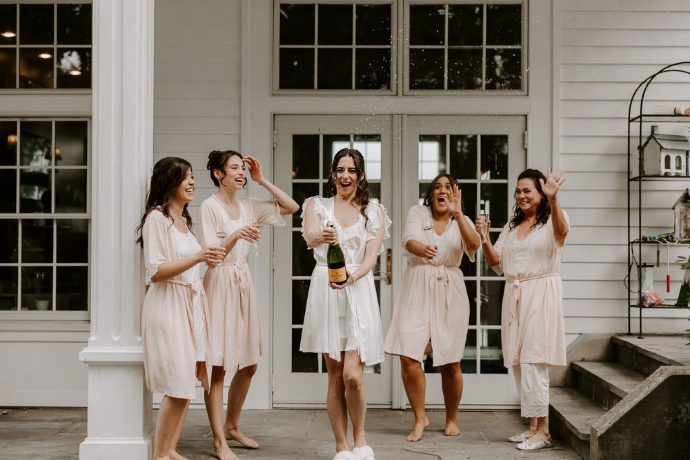 Bride in white robe popping champagne with bridesmaids on porch steps.