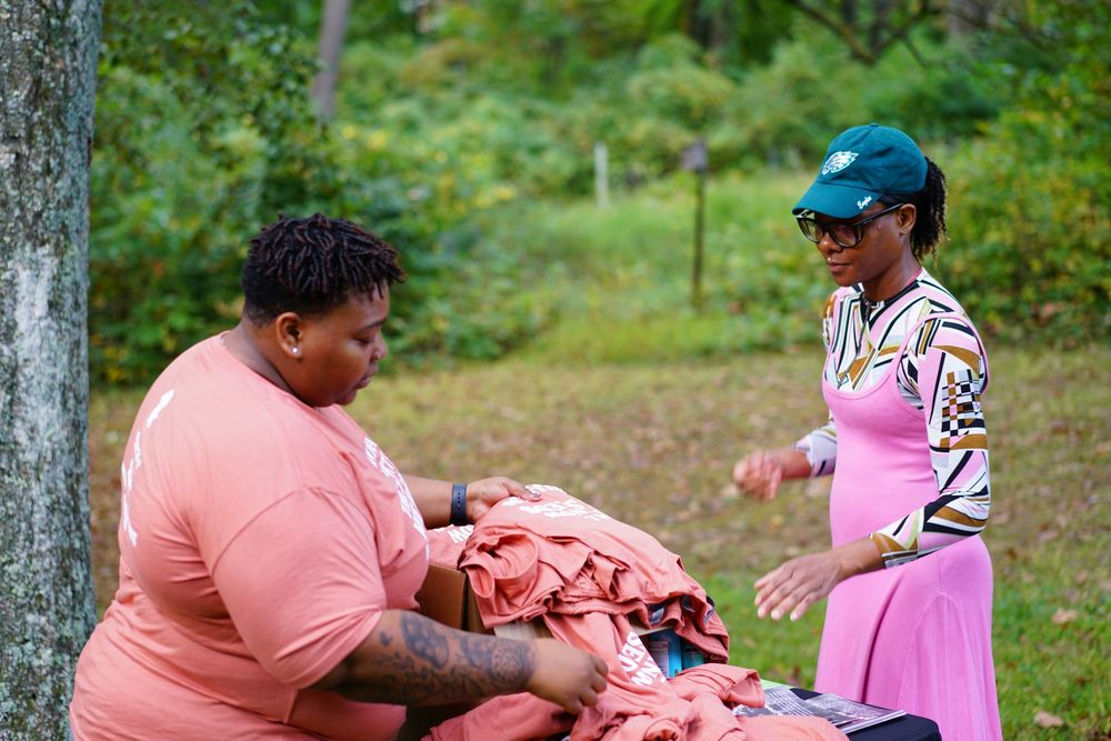 Two women outdoors exchange folded orange shirts near a tree in a grassy wooded area.