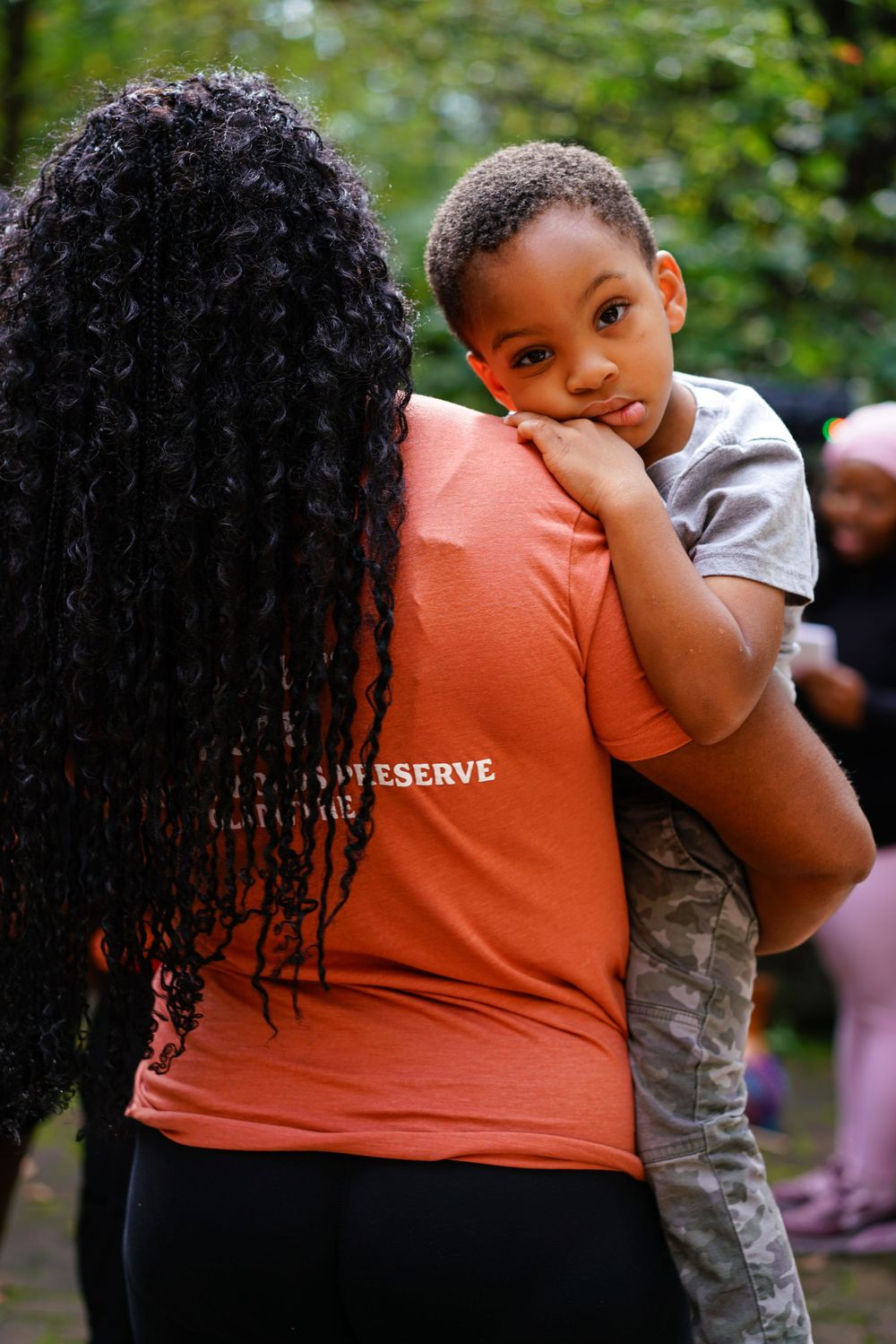 Woman in coral t-shirt holds a young boy on her hip outdoors at a community event.