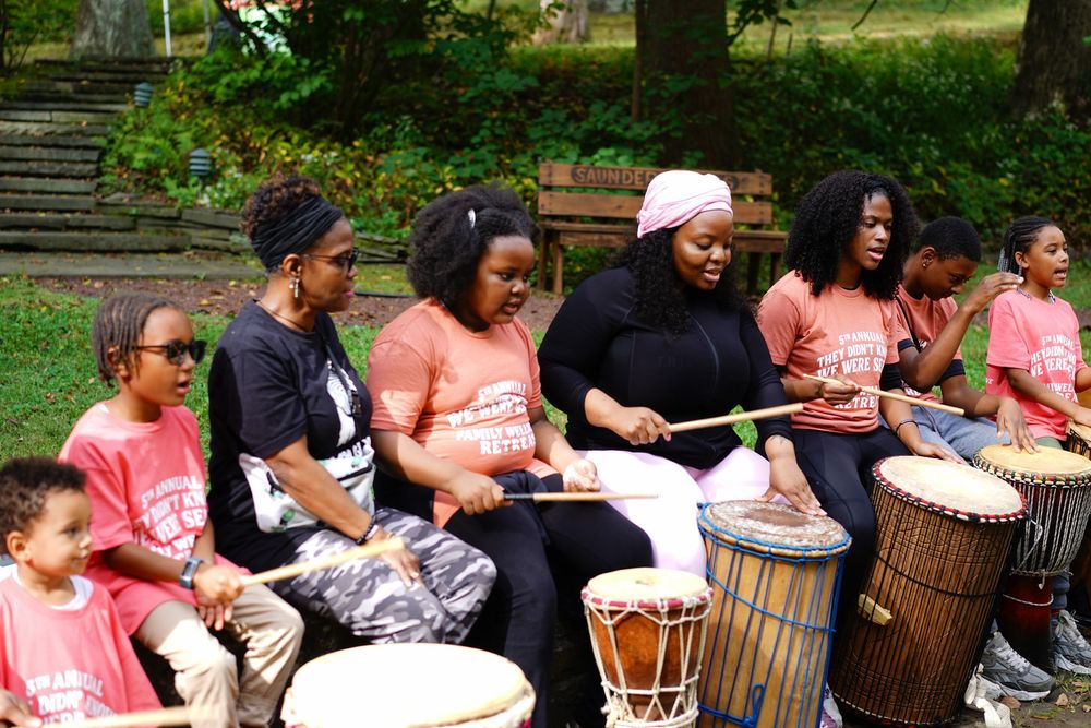 Group of people playing djembe drums outdoors in a park during a cultural drumming session.