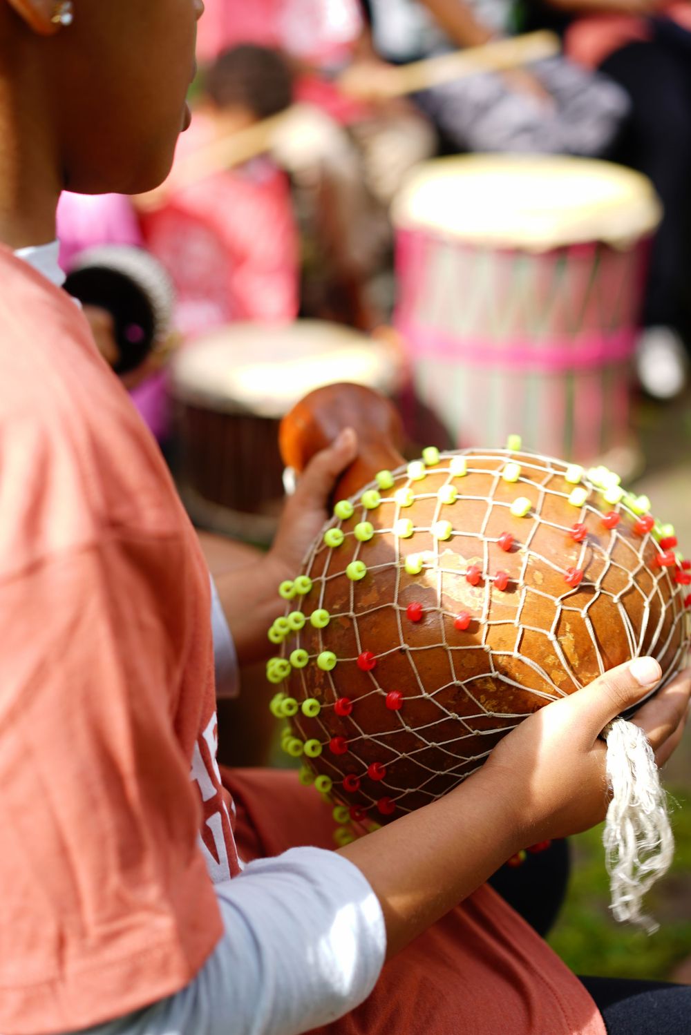 Person in orange shirt holds a traditional African shekere percussion instrument at a drum circle.