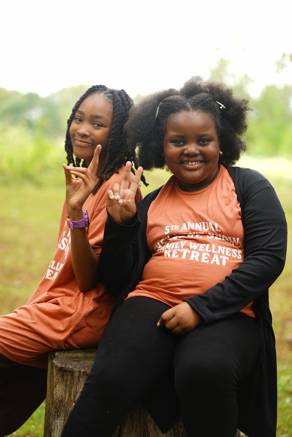 Two girls in orange wellness retreat shirts smile and pose outdoors, making peace signs near a wooden post.