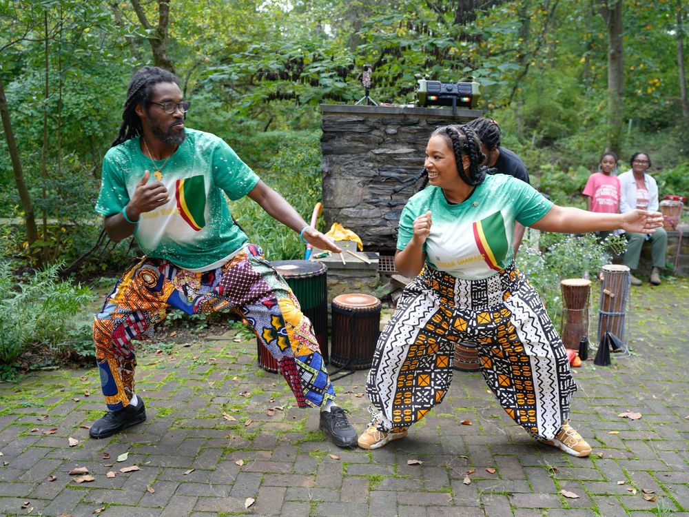 Two performers in colorful African attire dance energetically on a patio surrounded by lush greenery.