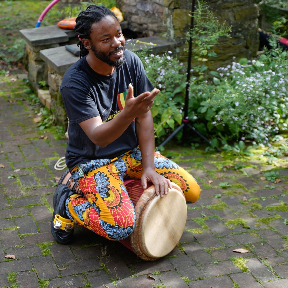 Smiling man in colorful African print pants plays a djembe drum outdoors on a stone patio.