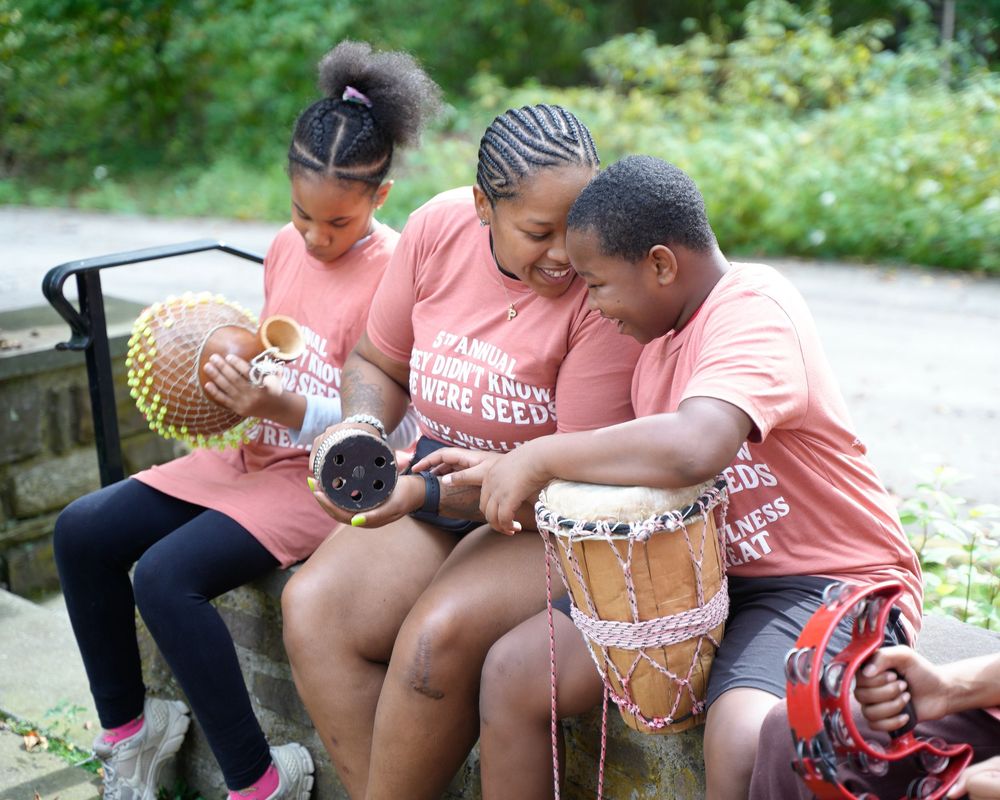 Three children in matching pink shirts play traditional African drums outdoors in a lush green setting.
