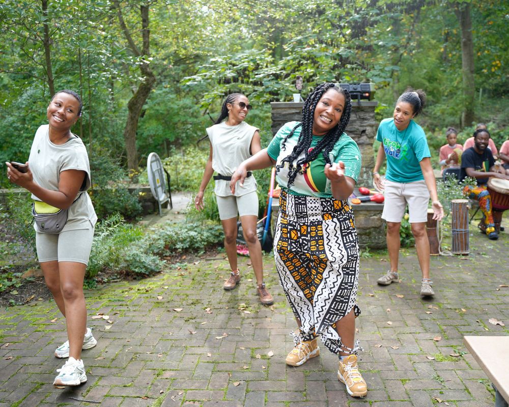Young women dancing and celebrating outdoors on a brick patio surrounded by lush green trees.