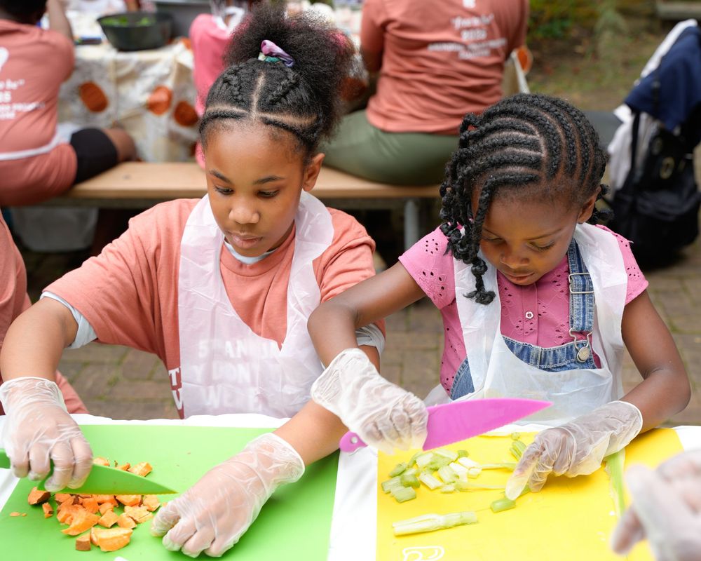 Two young girls wearing gloves chop colorful vegetables on cutting boards at a kids cooking activity.