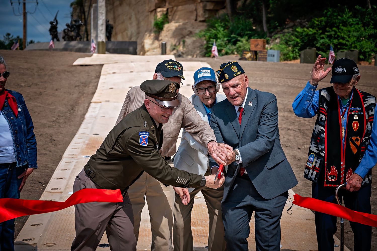 Veterans and officials cut a red ribbon at an outdoor ceremony, wearing military uniforms and caps at a historic stone site.