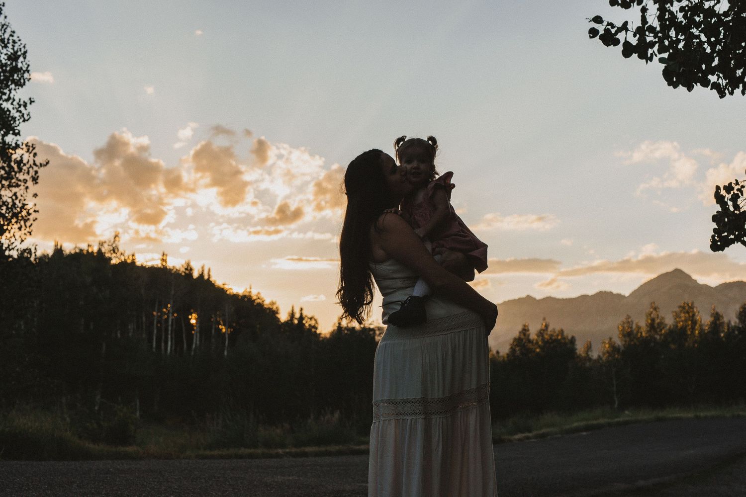 A mother kisses her baby at sunset with mountain silhouettes and golden sky in the background.