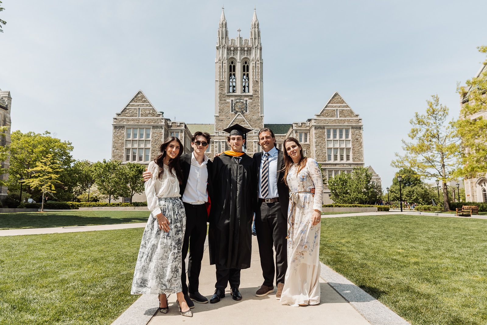 Graduate in cap and gown poses with four family members in front of a Gothic university building on a sunny day.