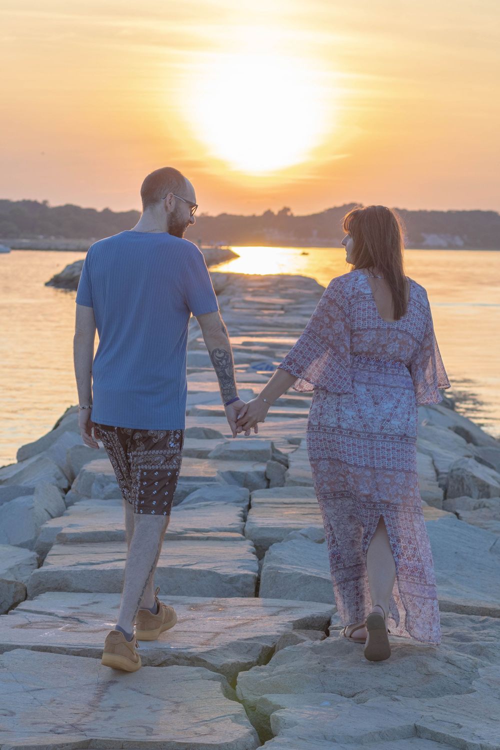 Couple holding hands walking on rocky pier toward golden sunset over calm ocean waters.