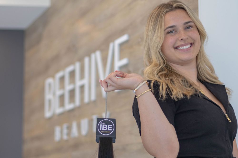Woman smiling and holding keys in front of Beehive Beauty salon wooden sign.