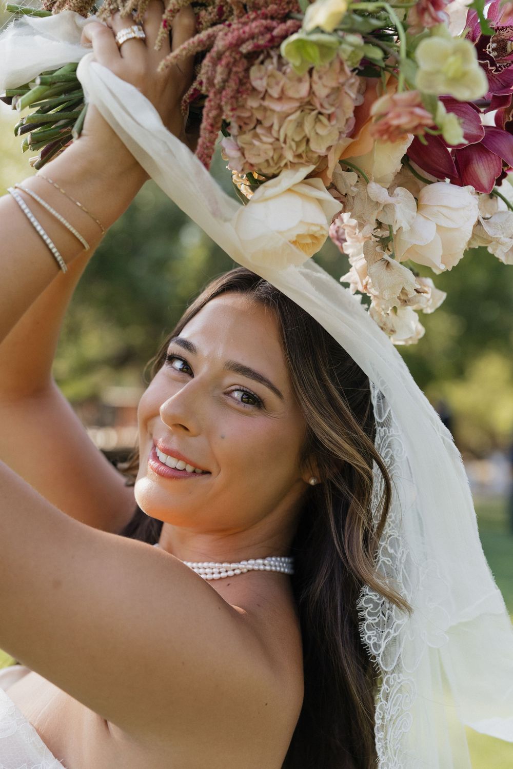 Smiling bride holds floral bouquet overhead, wearing pearl choker, veil, and bracelet on sunny outdoor wedding day.