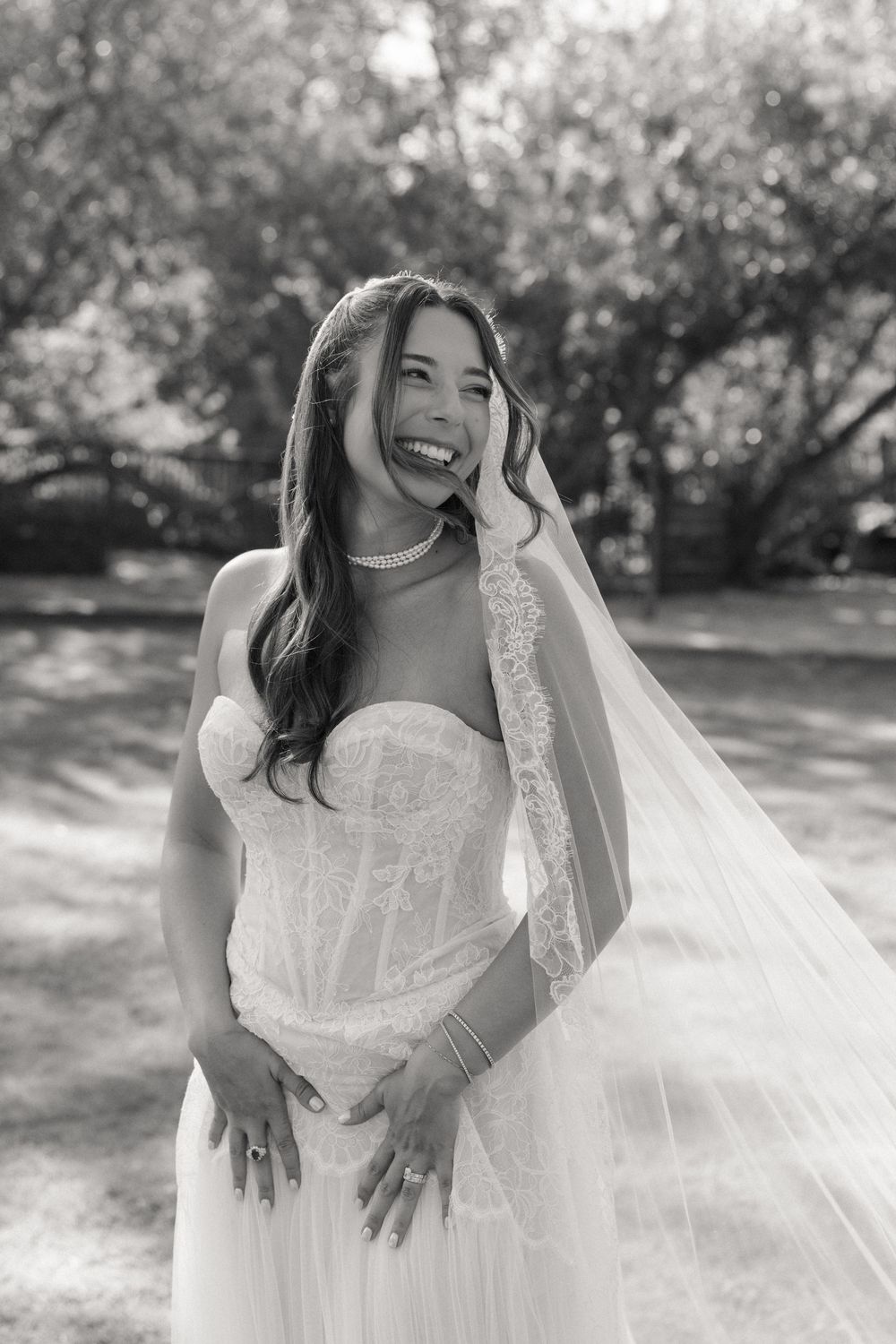 Black and white photo of a laughing bride in a strapless lace gown and veil posing outdoors.