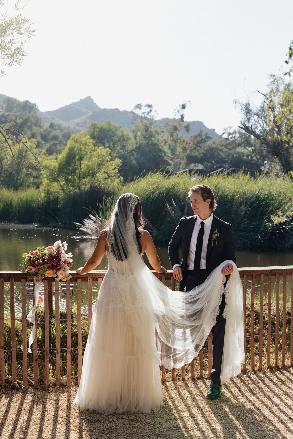 Bride and groom hold hands on a wooden deck overlooking a serene lake surrounded by lush greenery and mountains.