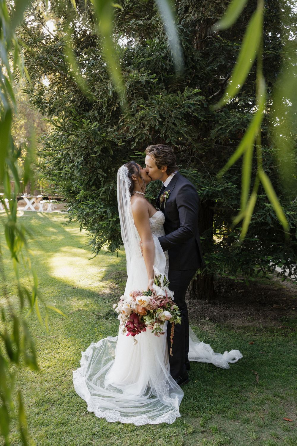 Bride and groom sharing a kiss in a lush garden, bride wearing a flowing white gown with veil holding a colorful bouquet.