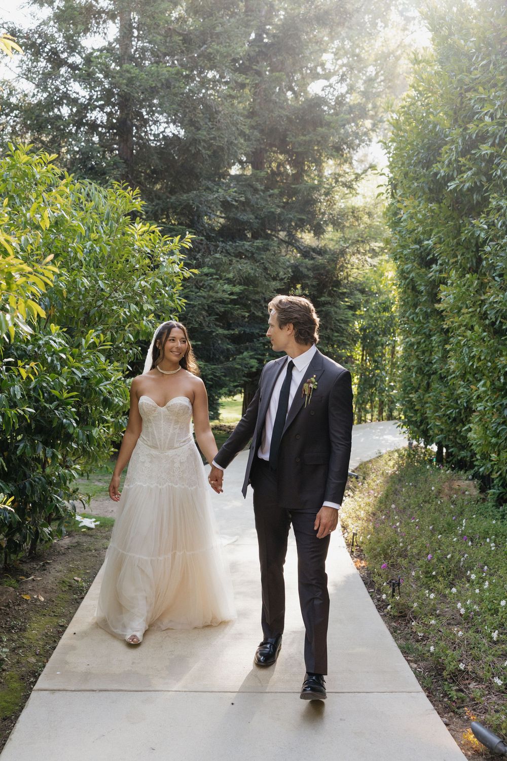 Bride and groom walk hand-in-hand along a sunlit garden path surrounded by lush green trees.