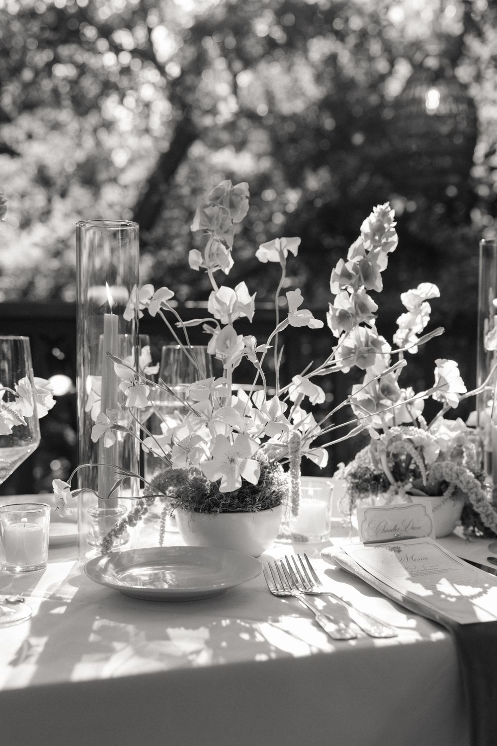 Black and white outdoor wedding table setting with floral centerpiece, glassware, and dappled sunlight through trees.