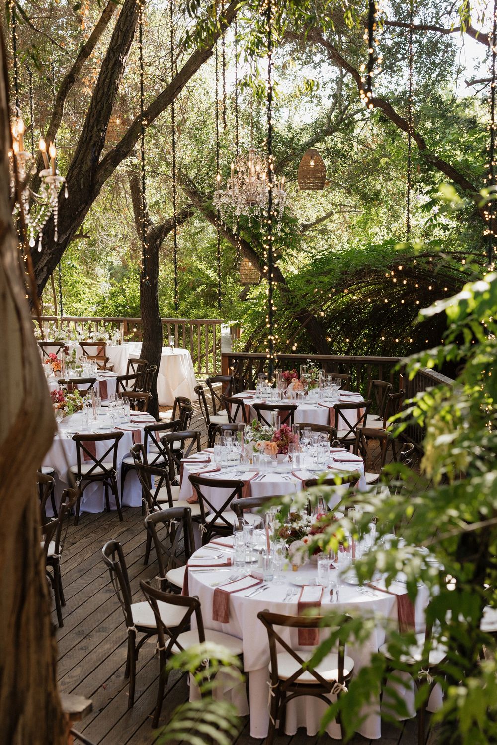Elegant outdoor wedding reception on a wooded deck with round tables, string lights, and lush greenery.
