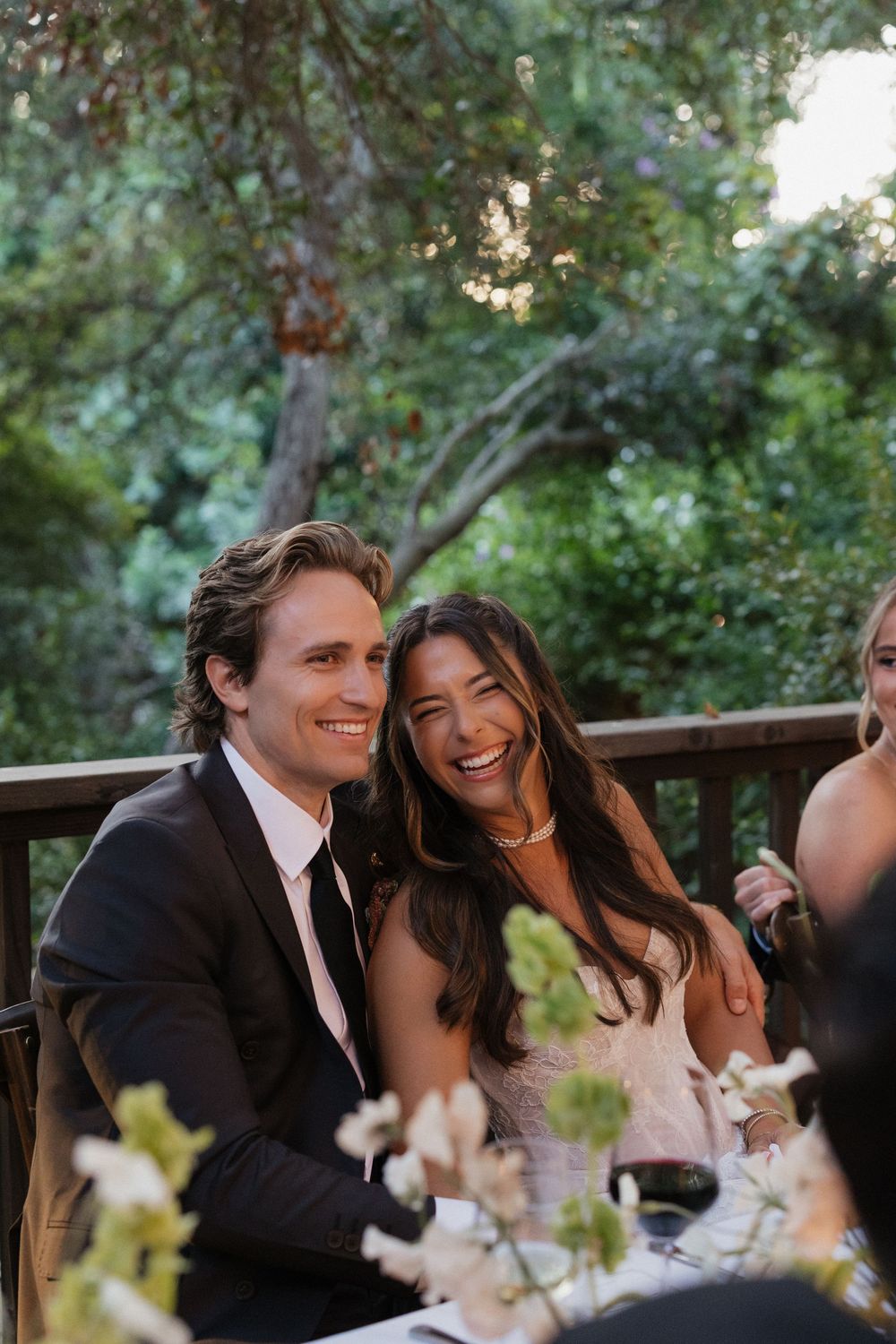 A smiling couple in formal attire sits together at an outdoor wedding reception surrounded by lush greenery.