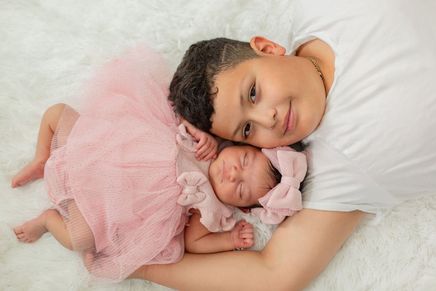 A young girl in a pink tutu lies on white fur beside a sleeping newborn baby in a pink bow headband.