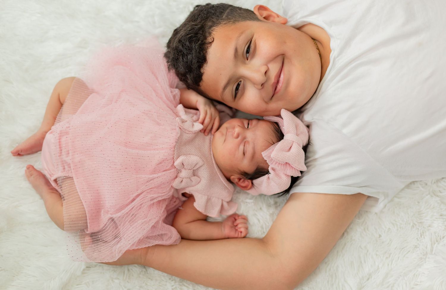 Older child smiling while lying next to sleeping newborn baby in pink dress and bow headband on white background.