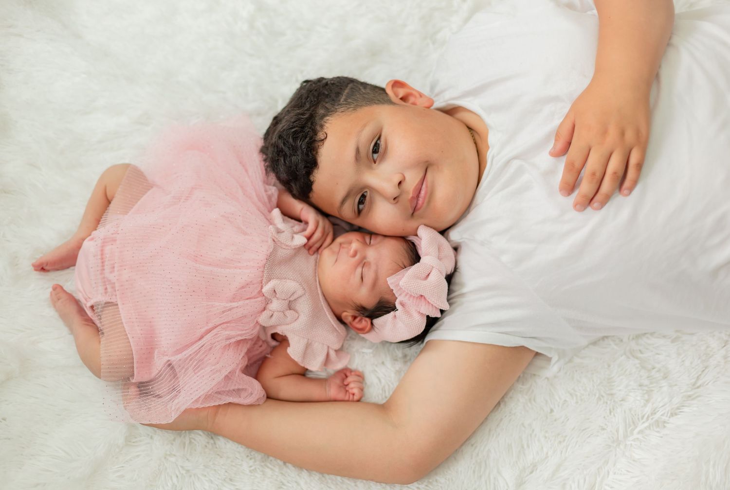 A young child tenderly holds a newborn baby dressed in pink on a white fluffy blanket.