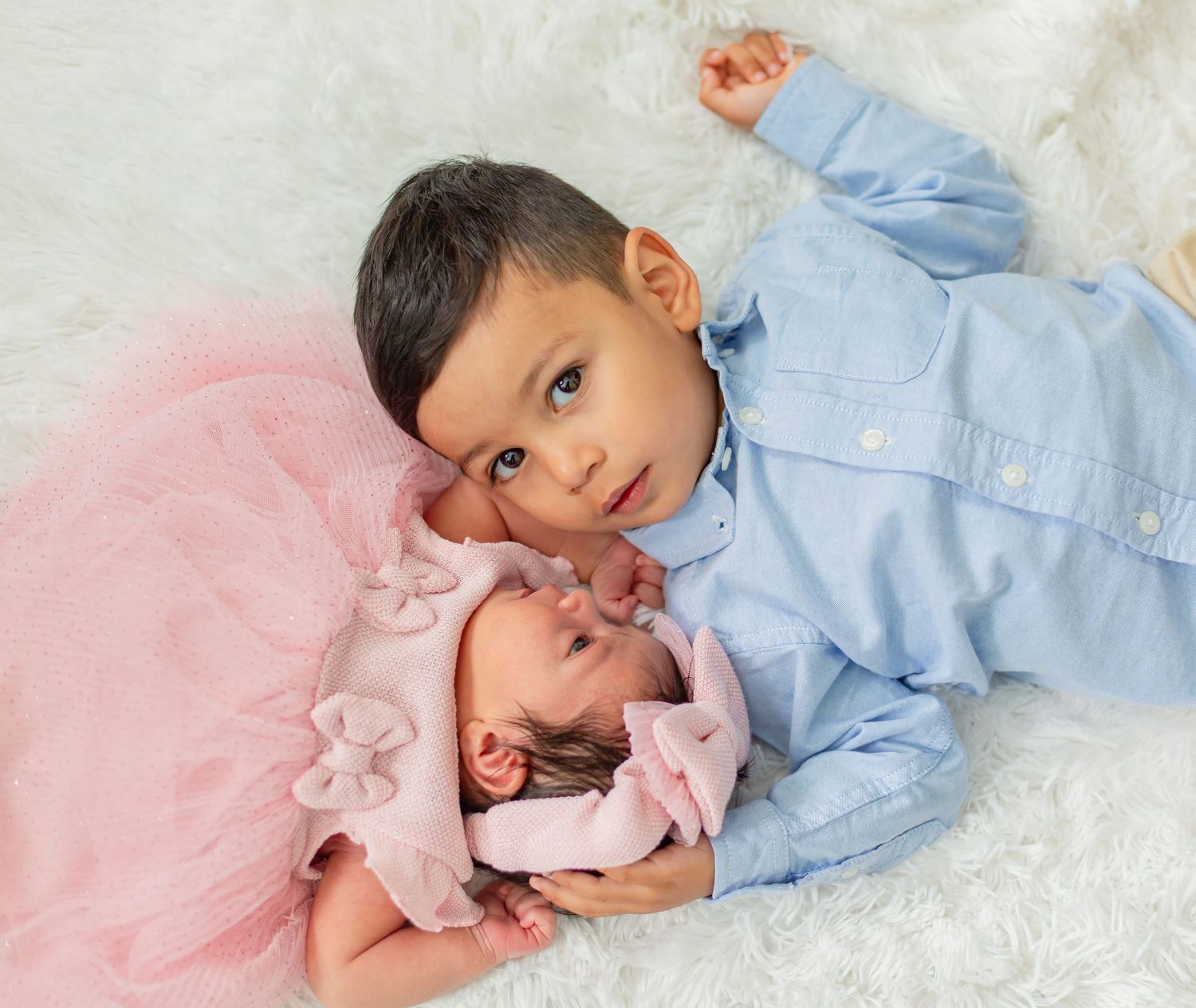 A young toddler boy in a blue shirt cuddles with a newborn baby girl in a pink tutu on a white fur blanket.