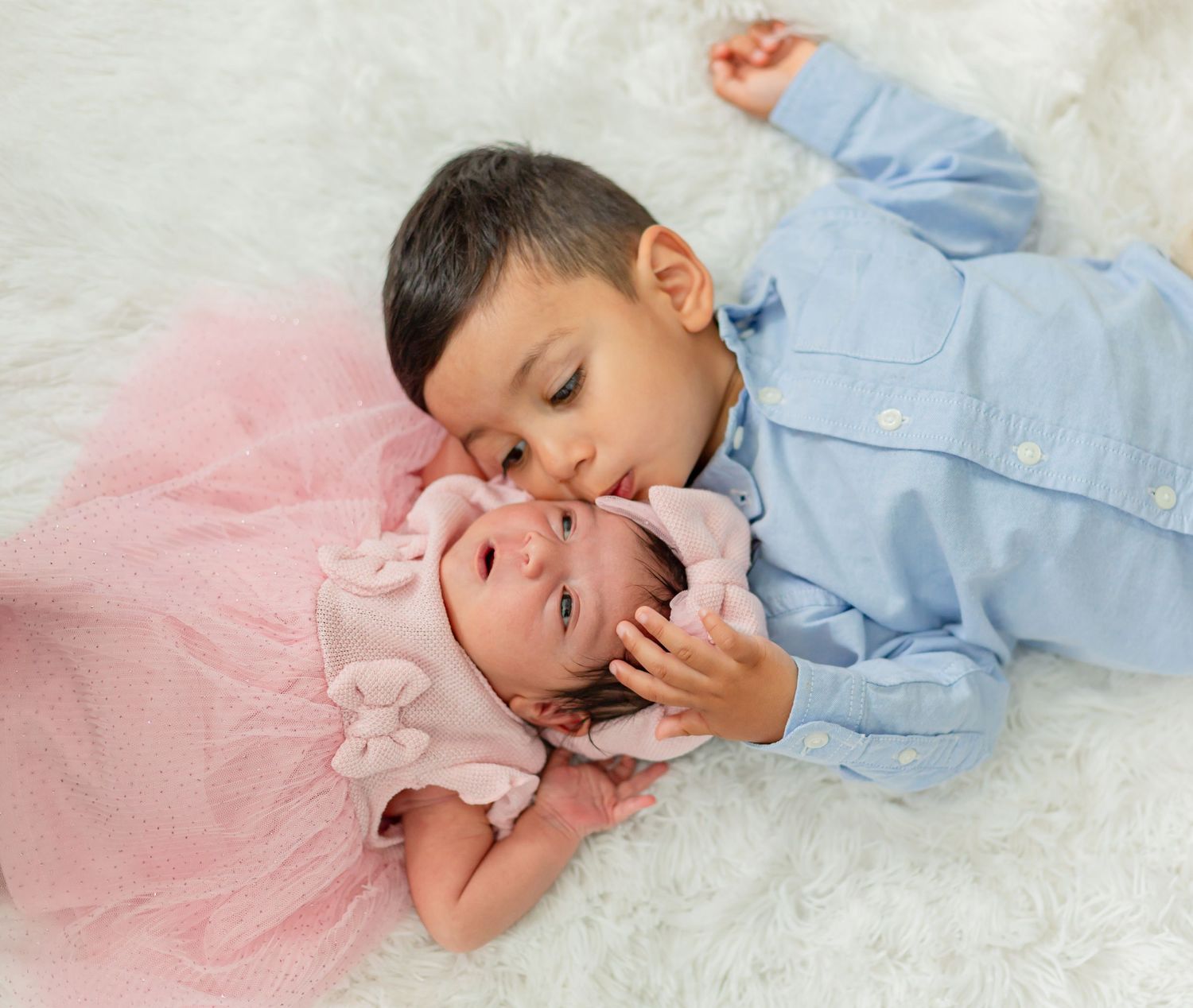 Young boy in blue shirt kisses newborn baby girl in pink tutu dress, both lying on white fluffy rug.