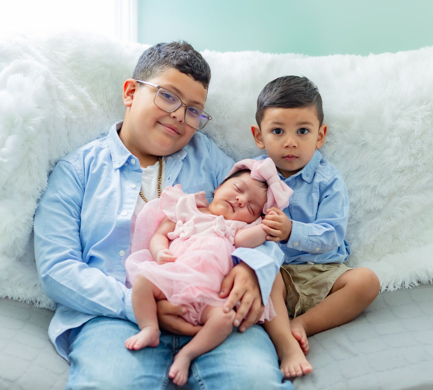 Two boys in blue shirts pose with a newborn baby girl in a pink bow on a white fluffy couch.