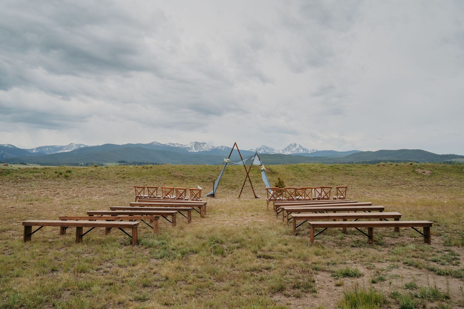 Outdoor wedding ceremony setup with wooden benches and triangular arch in a mountain meadow under cloudy skies.