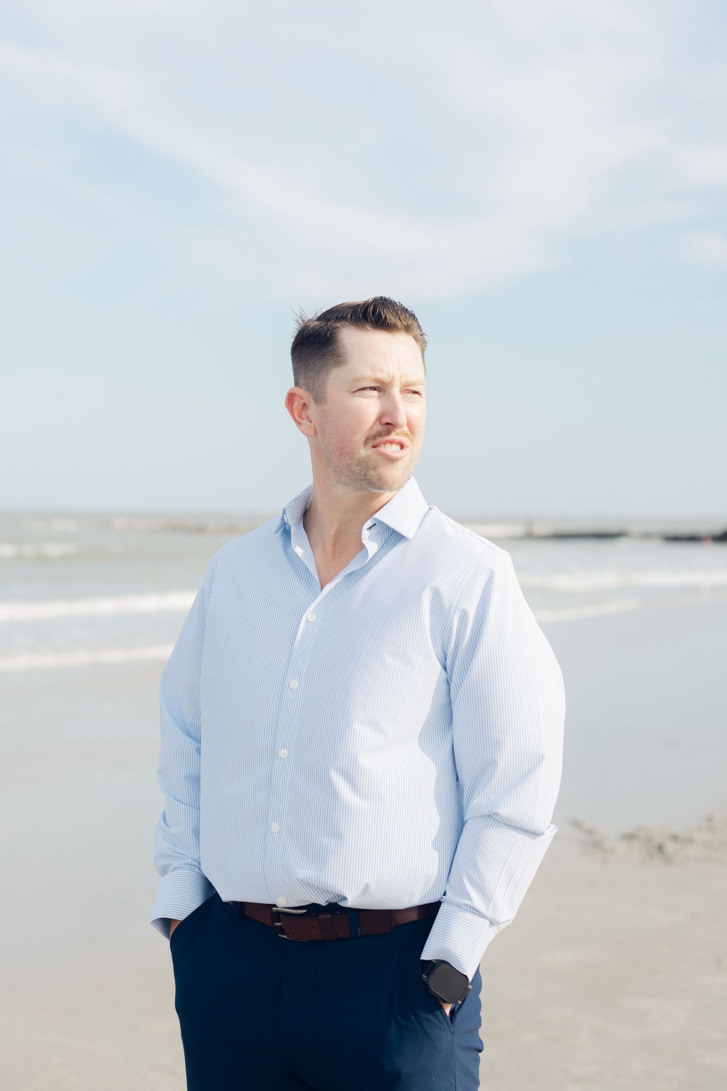 A man in a light blue dress shirt and navy pants stands on a sunny beach with hands in pockets, smiling.