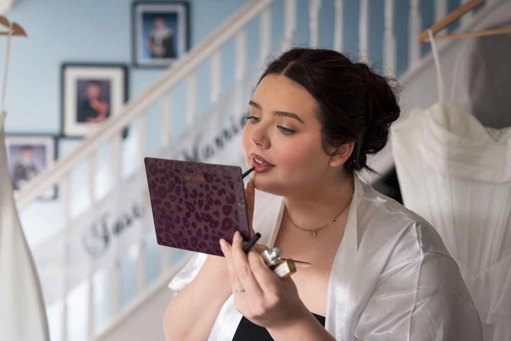 Someone in a white robe examines a small purple book while sitting near a staircase with framed photos and a dress.