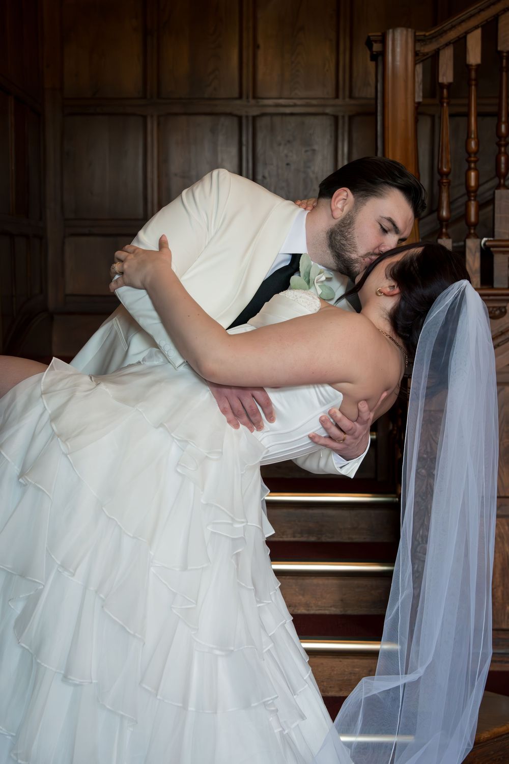 A romantic wedding couple embraces on wooden stairs in a dramatic dip pose with flowing white dress and veil.