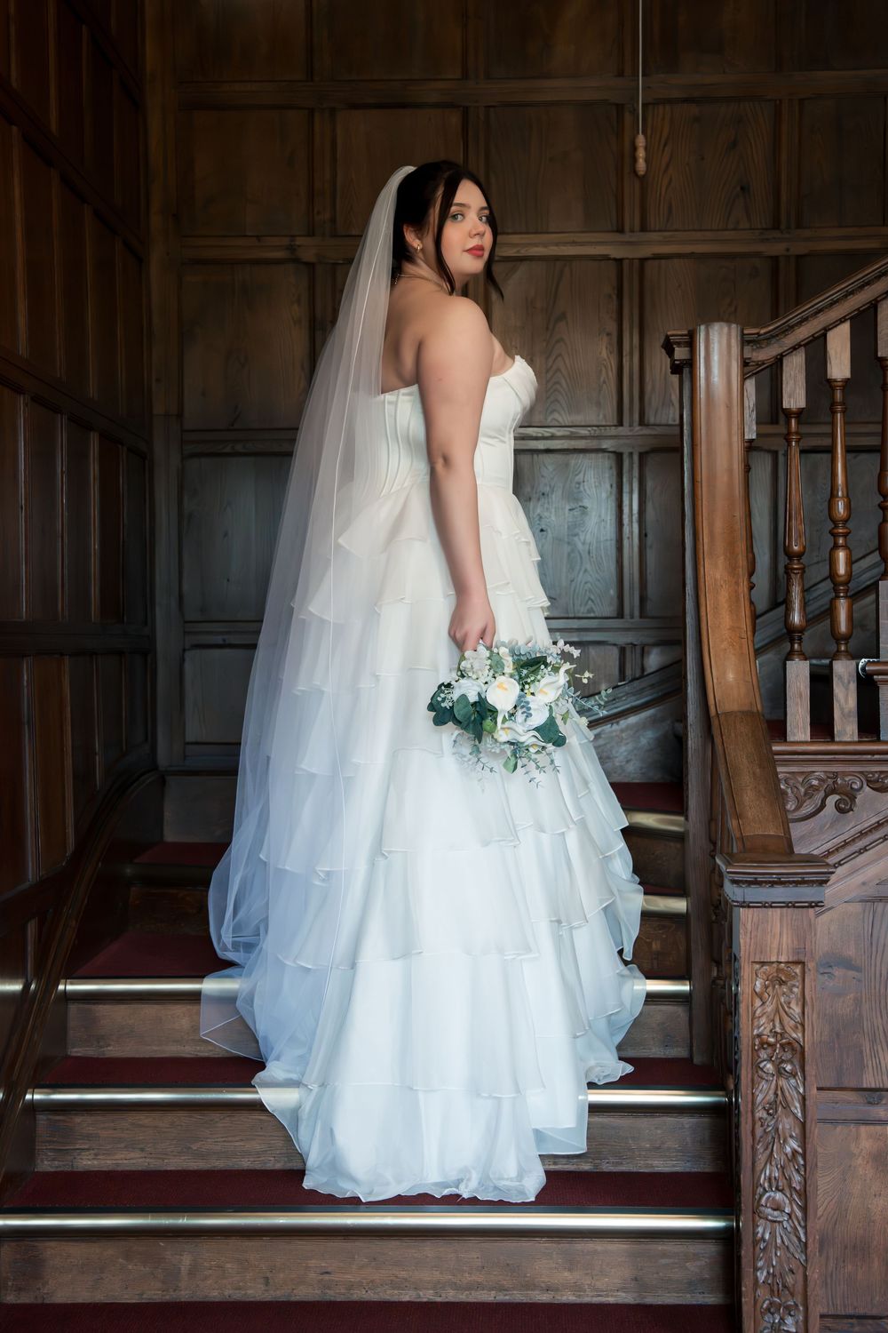 A bride in a white wedding gown and veil stands on ornate wooden stairs in a vintage interior setting.