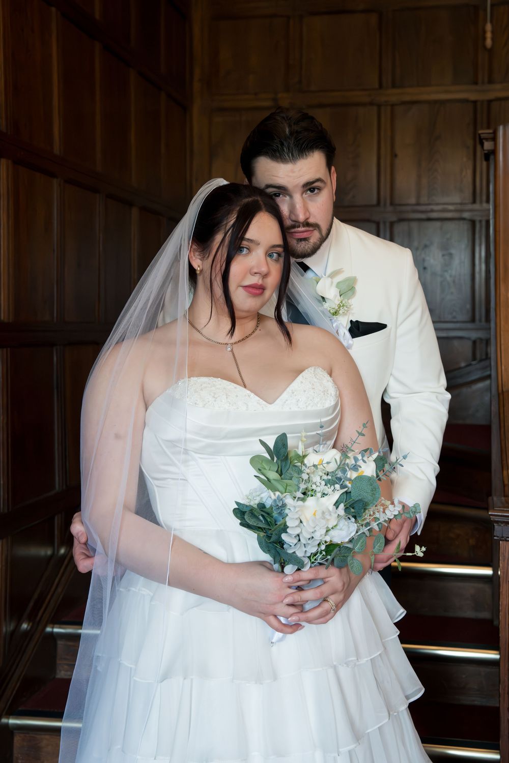A romantic wedding couple embraces in a wood-paneled room with white floral bouquet and elegant bridal attire.