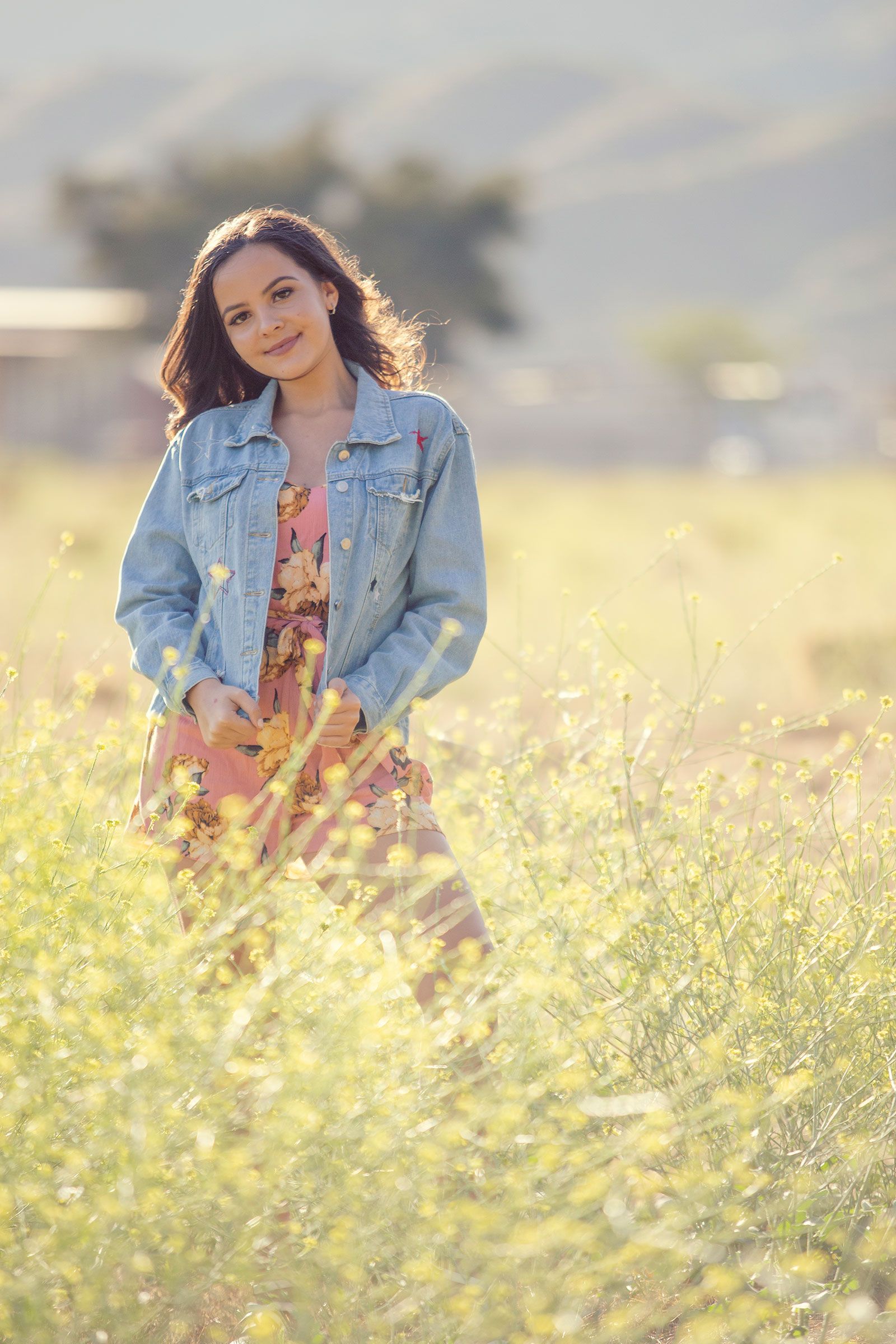 Smiling brunette teen in floral dress and denim jacket poses in a golden wildflower meadow at sunset.