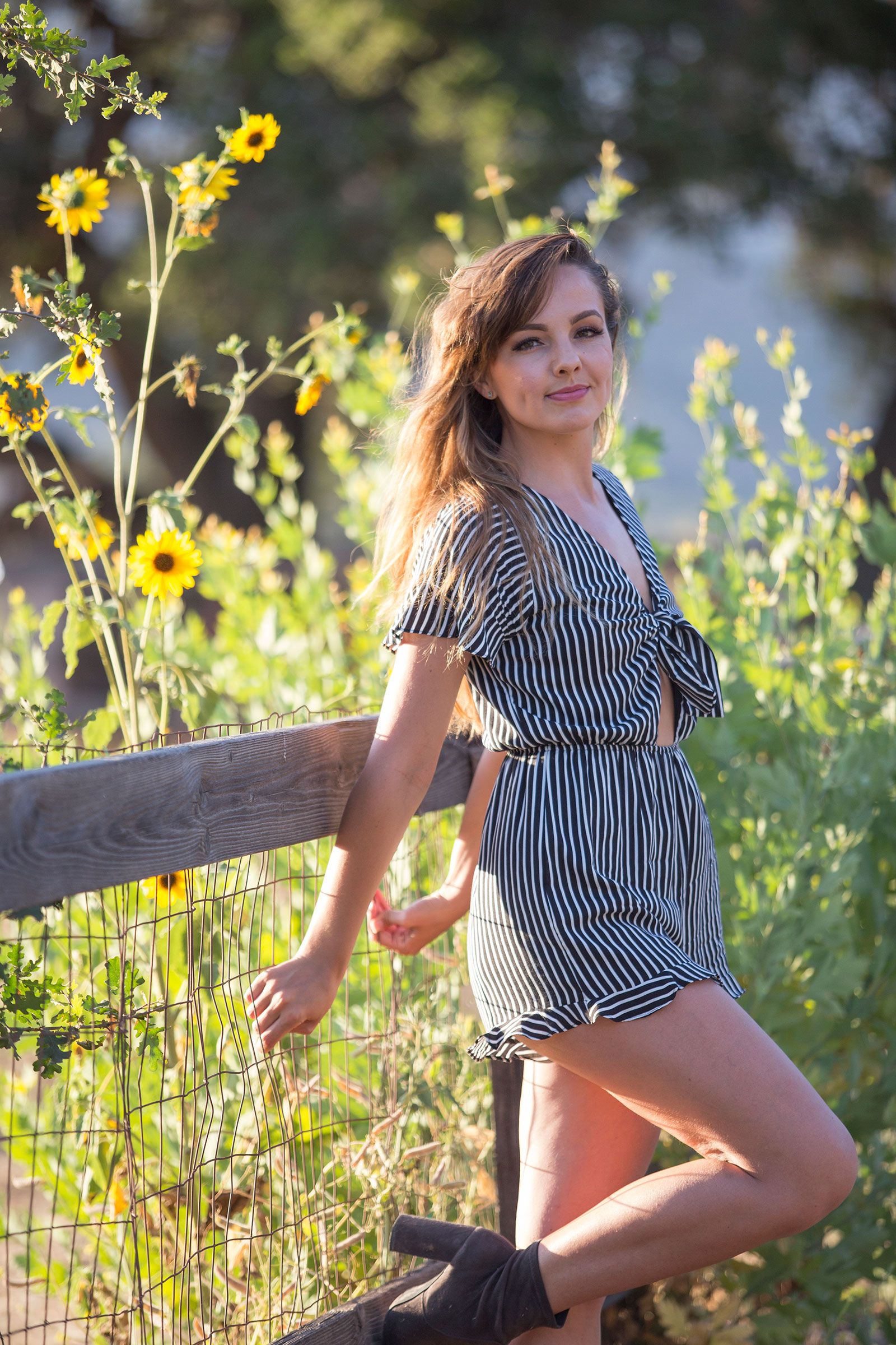 Smiling woman in striped romper leans against a wooden fence surrounded by bright yellow wildflowers.
