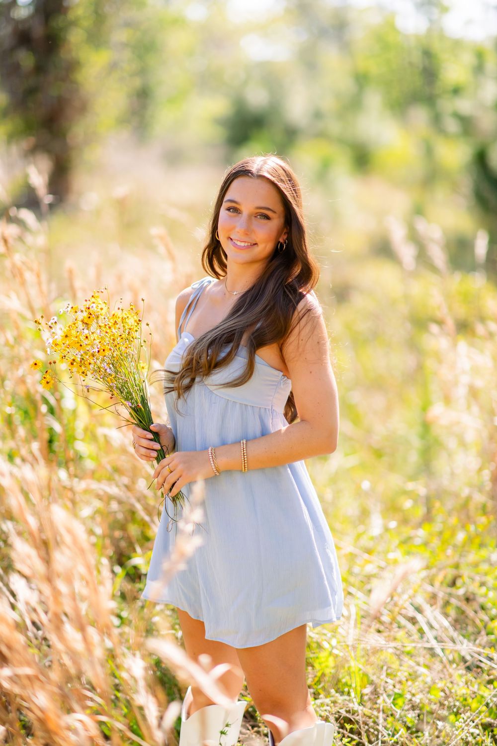 Young woman in blue dress holding yellow wildflowers, smiling in a sunlit field.
