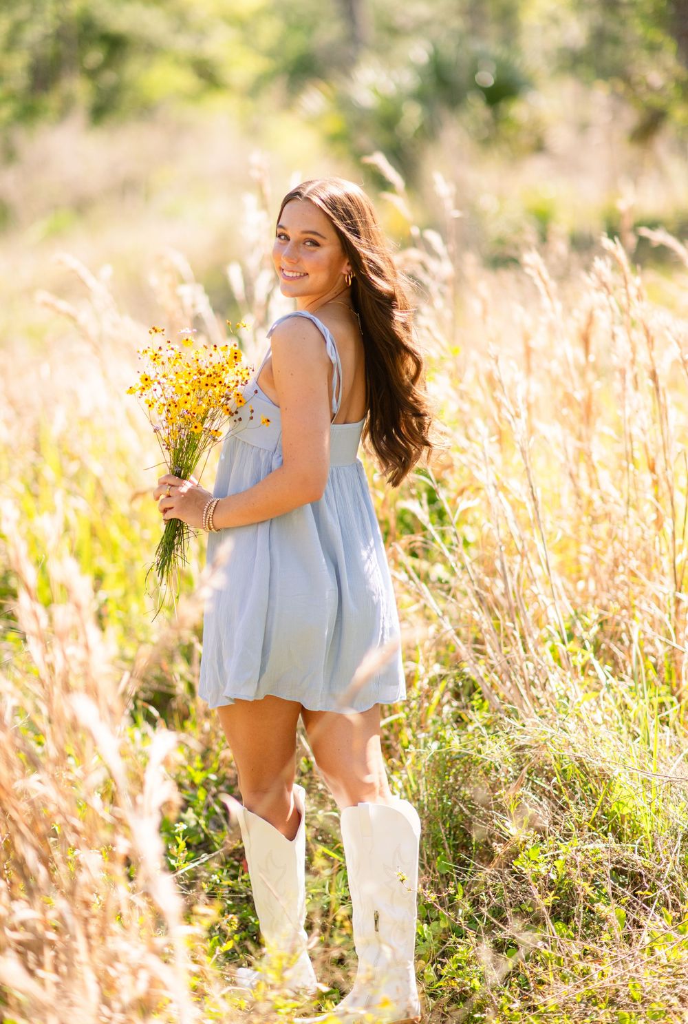 Smiling brunette teen in light blue dress and white cowboy boots holding yellow wildflowers in sunny field.