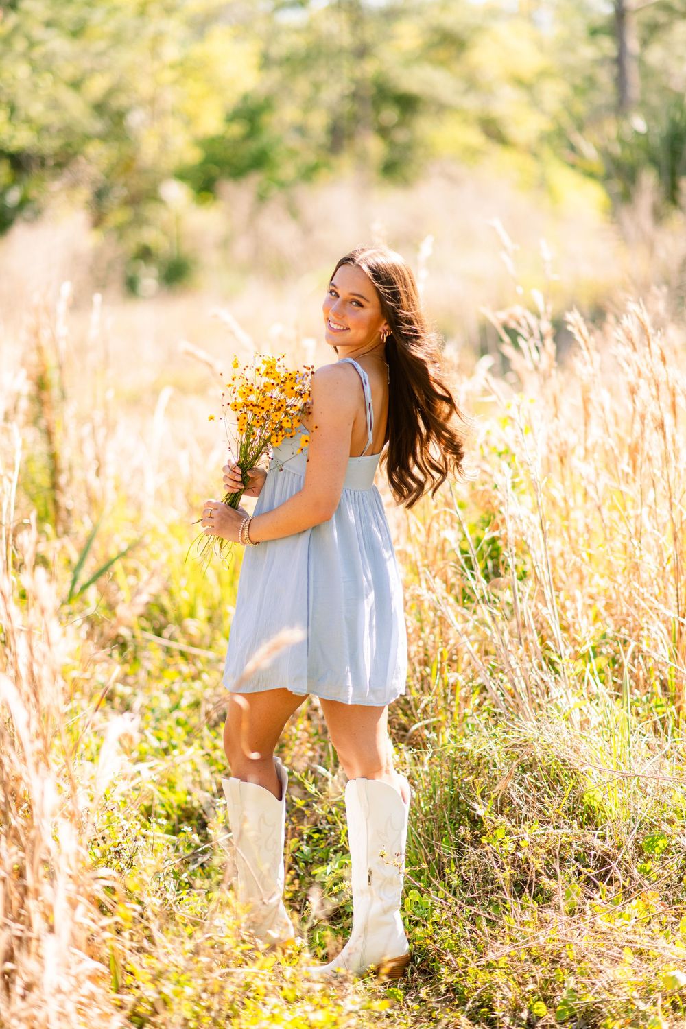 Smiling young woman in blue dress and white boots holding yellow wildflowers in field.