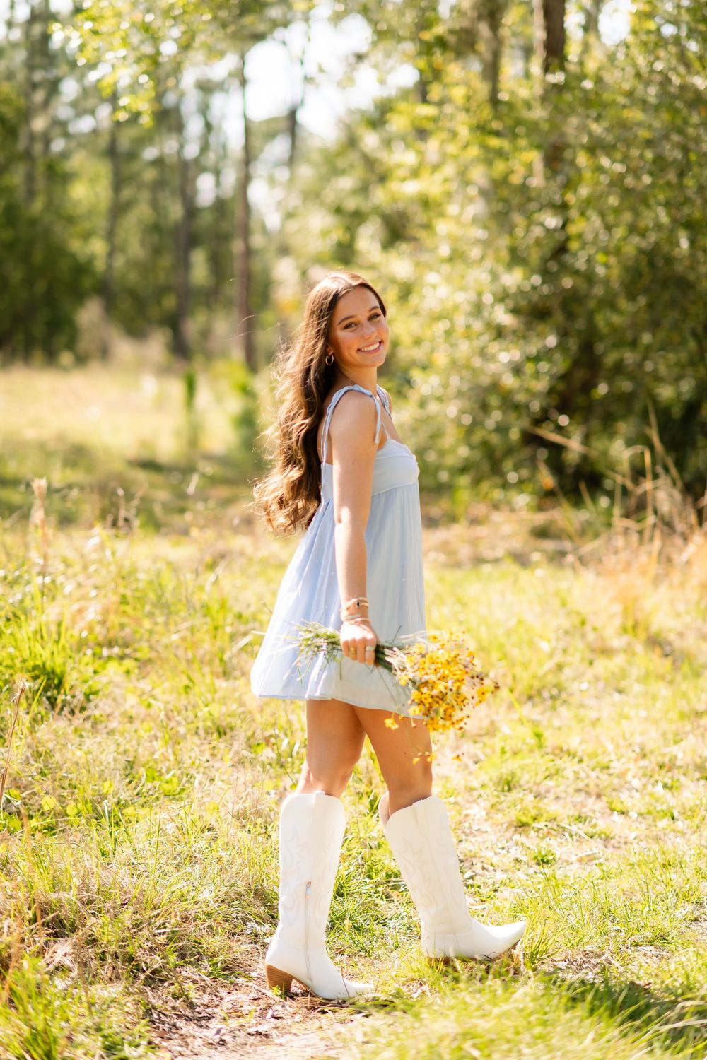Smiling brunette girl in blue dress and white cowboy boots holding yellow wildflowers in sunny forest path.