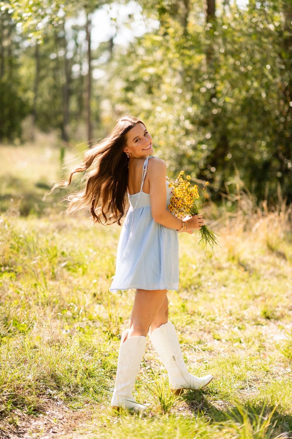 Smiling young woman in a light blue dress and white cowboy boots holding yellow wildflowers in a sunny forest.